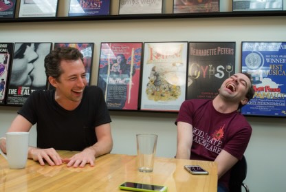 Two men laughing, one in a black shirt and one in a maroon t-shirt with "GOD HATE SHAKESPEARE" printed on it.