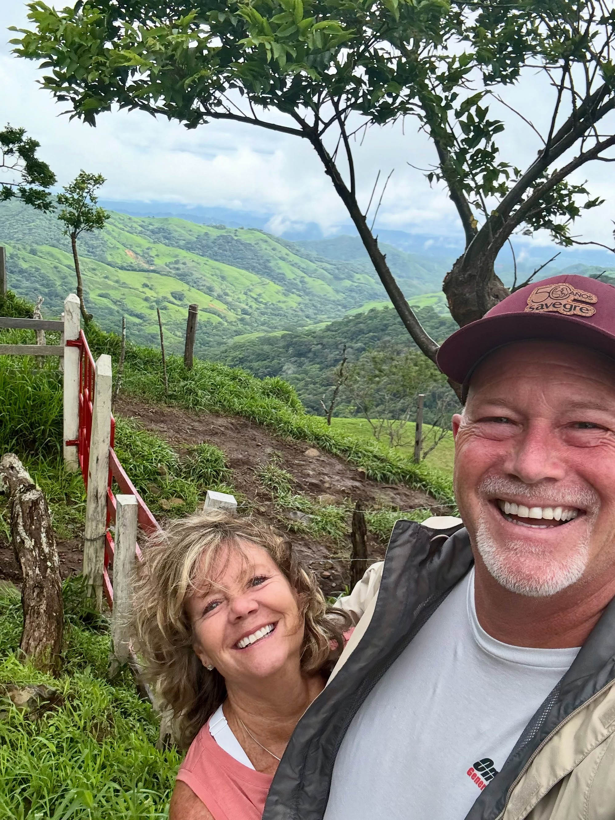 Selfie of a smiling man and woman with a mountainous, tree-filled landscape in the background.