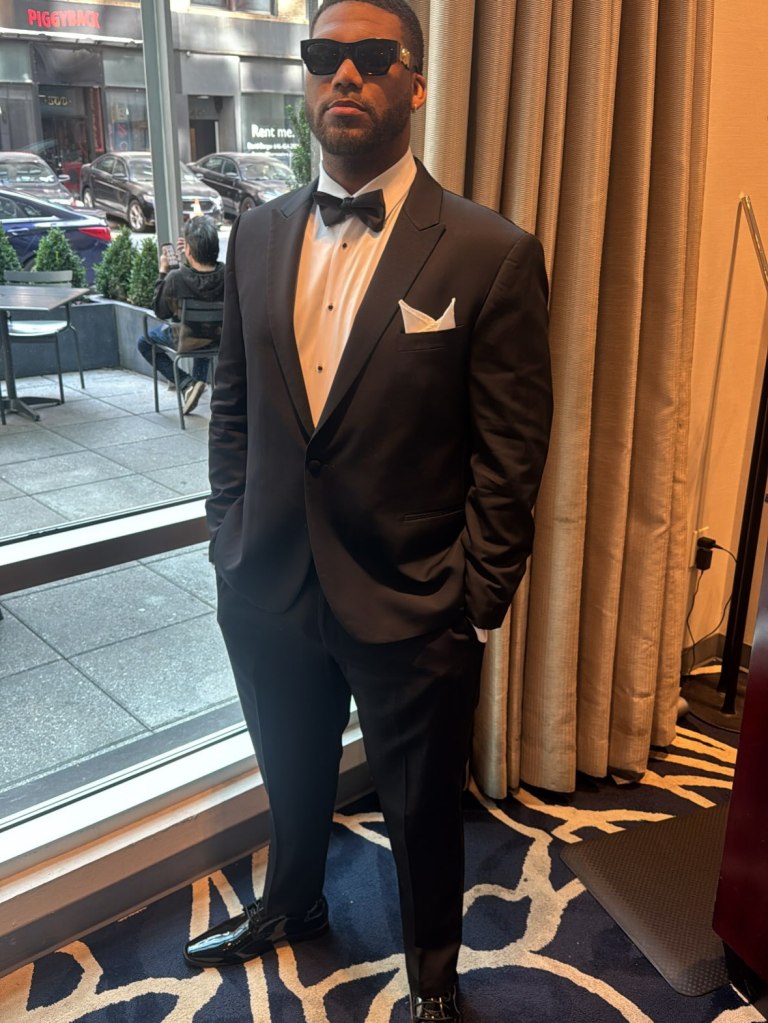 A man in a tuxedo and sunglasses stands with hands in pockets.