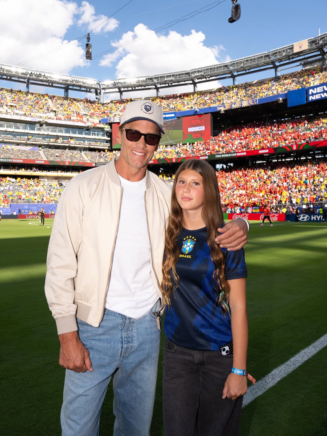 Tom Brady smiling with his daughter in a crowded soccer stadium.