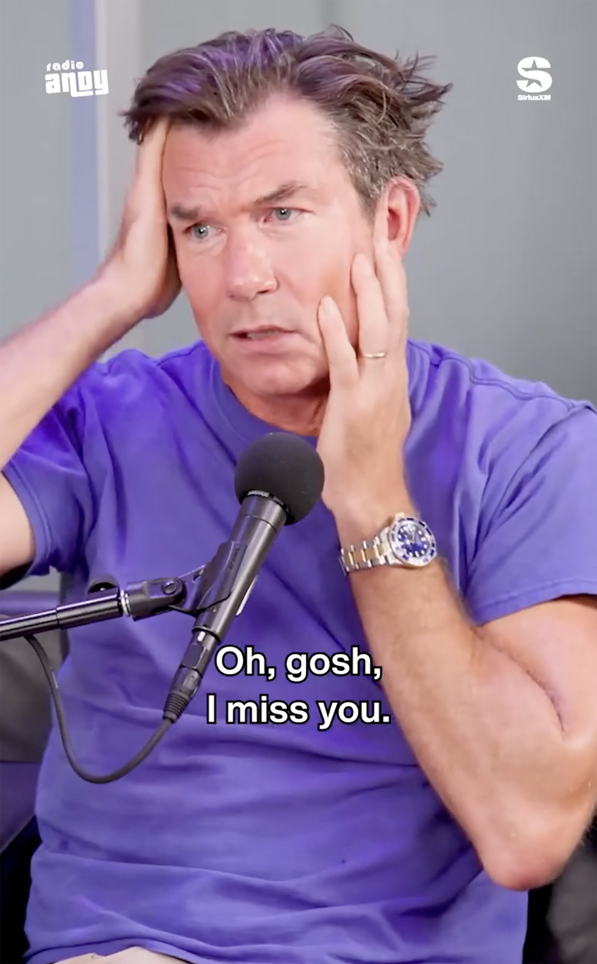 Jerry O'Connell holding his head in his hands, wearing a purple shirt, with a microphone in front of him.