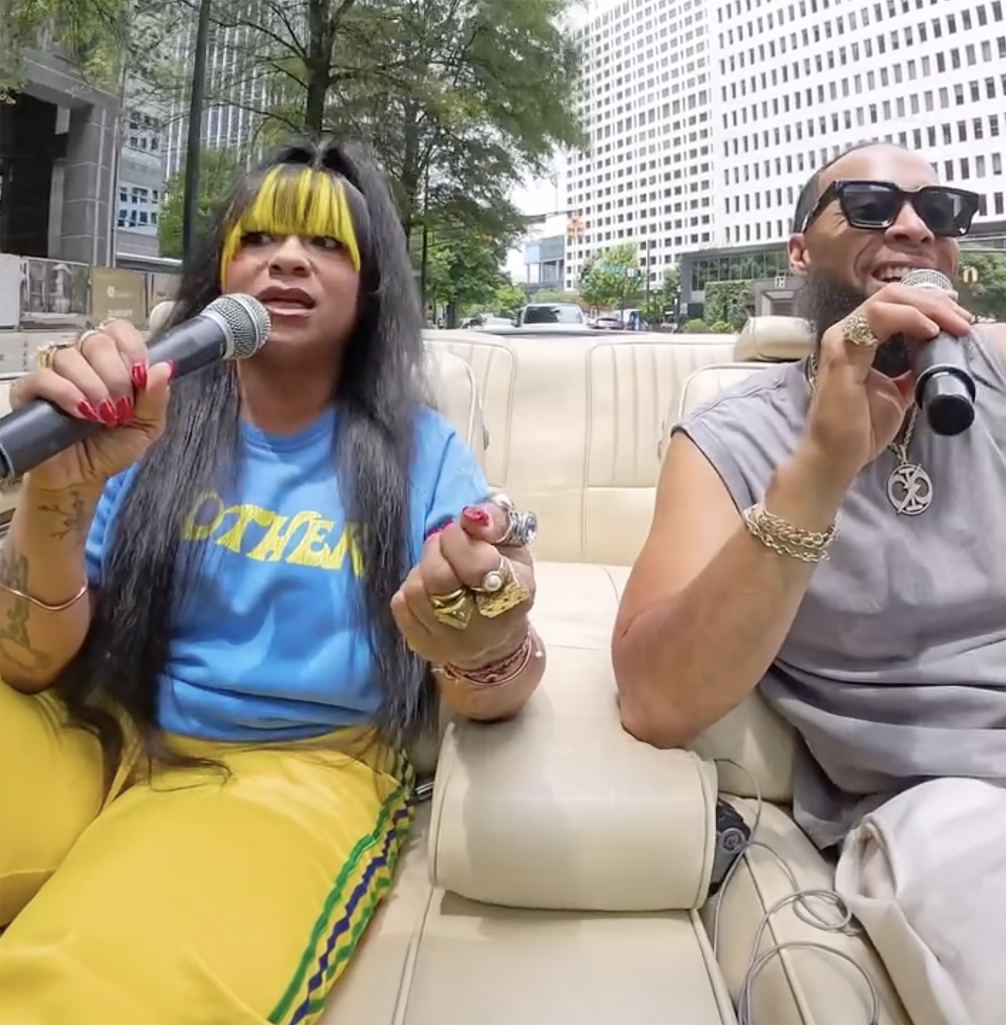 Nivea and a man sing karaoke in a white convertible, with city buildings and trees in the background.