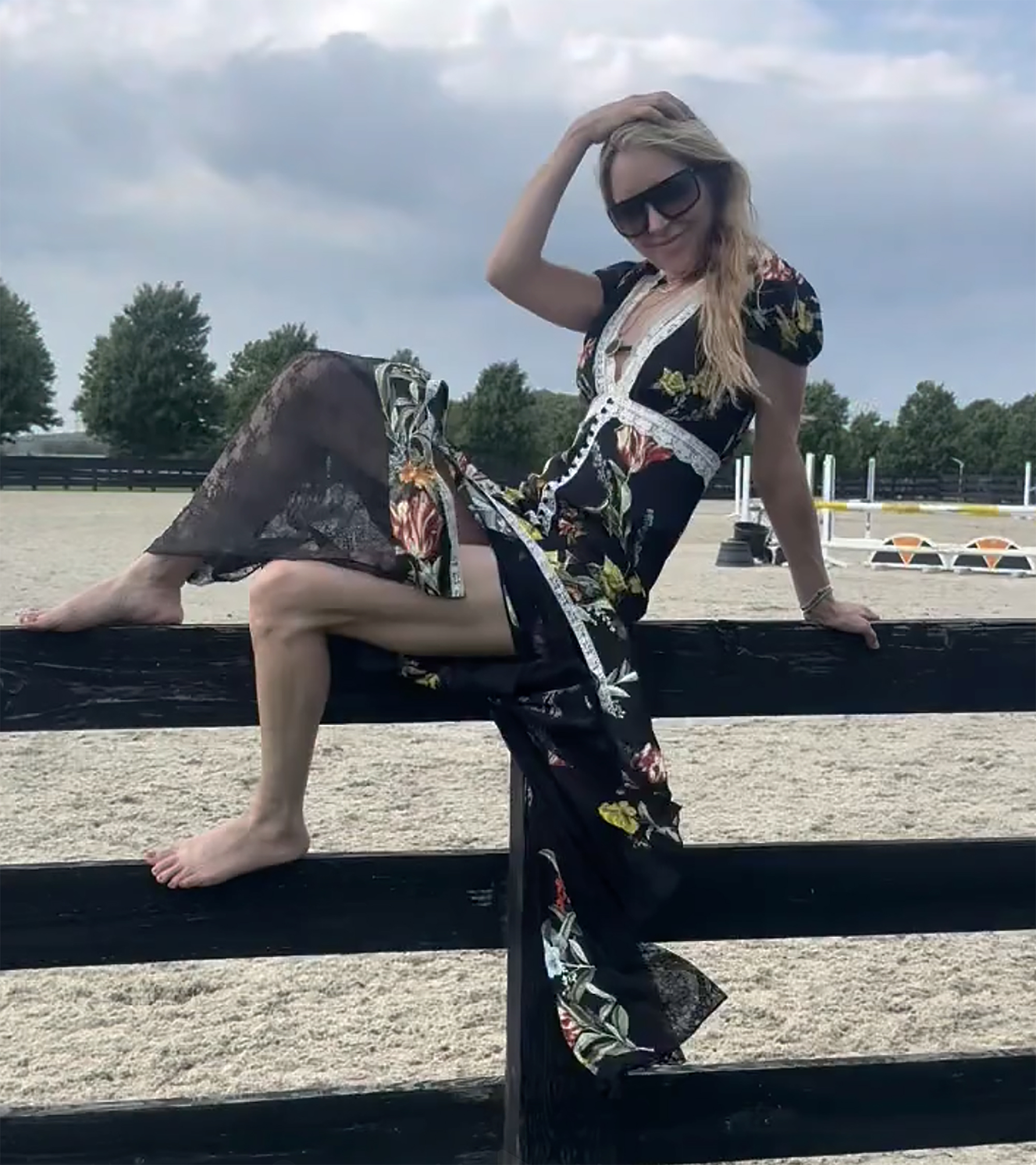 Jenny Mollen sitting barefoot on a black fence in a flowered dress and sunglasses.