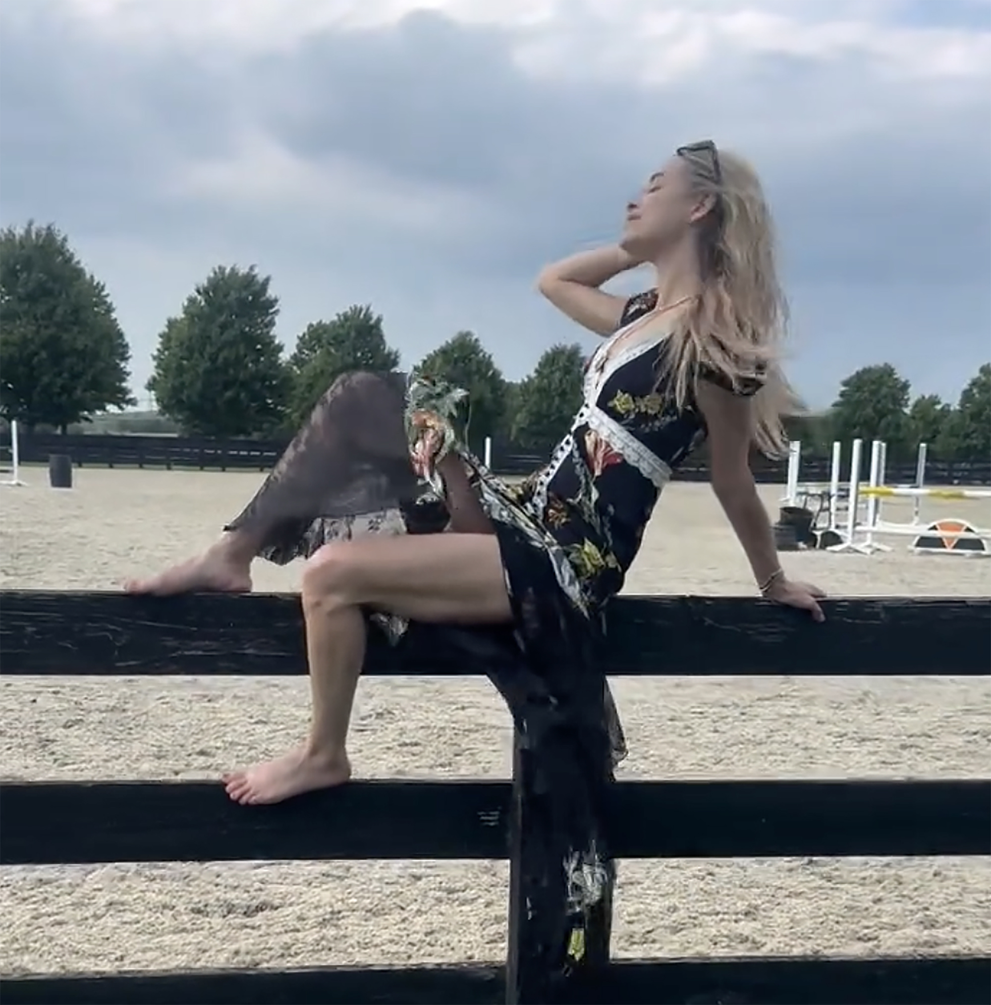 Jenny Mollen in a floral dress, barefoot on a fence at an equestrian training ground.