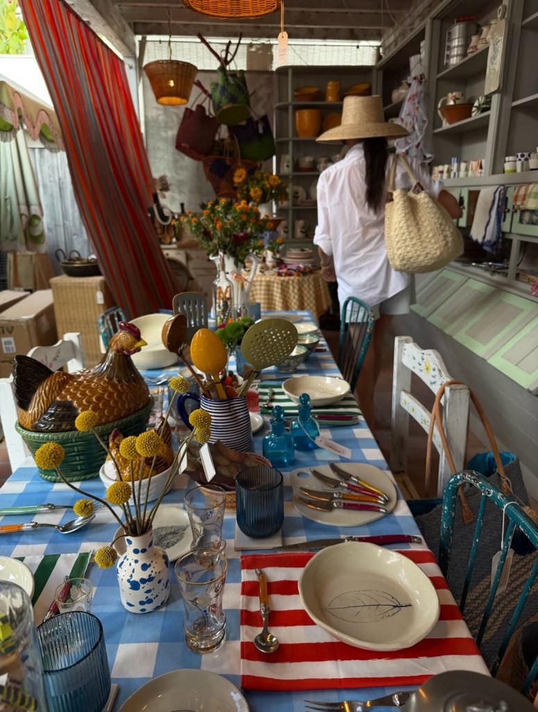 A woman in a sun hat and white shirt holding a straw bag stands in a rustic shop filled with colorful homewares, including a table set with checkered and striped tablecloths, ceramic dishes, and yellow floral arrangements.