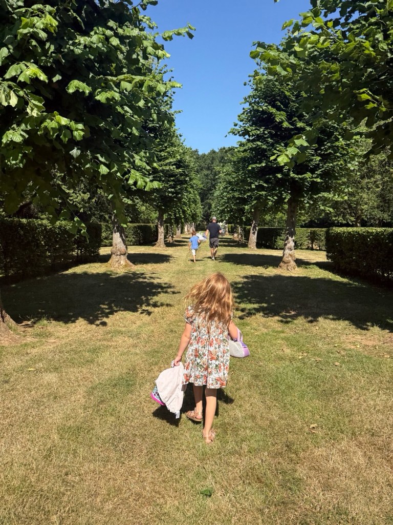 A little girl in a floral dress walks on grass toward a man and a boy, all viewed from behind, in an outdoor alley lined with trees and hedges.