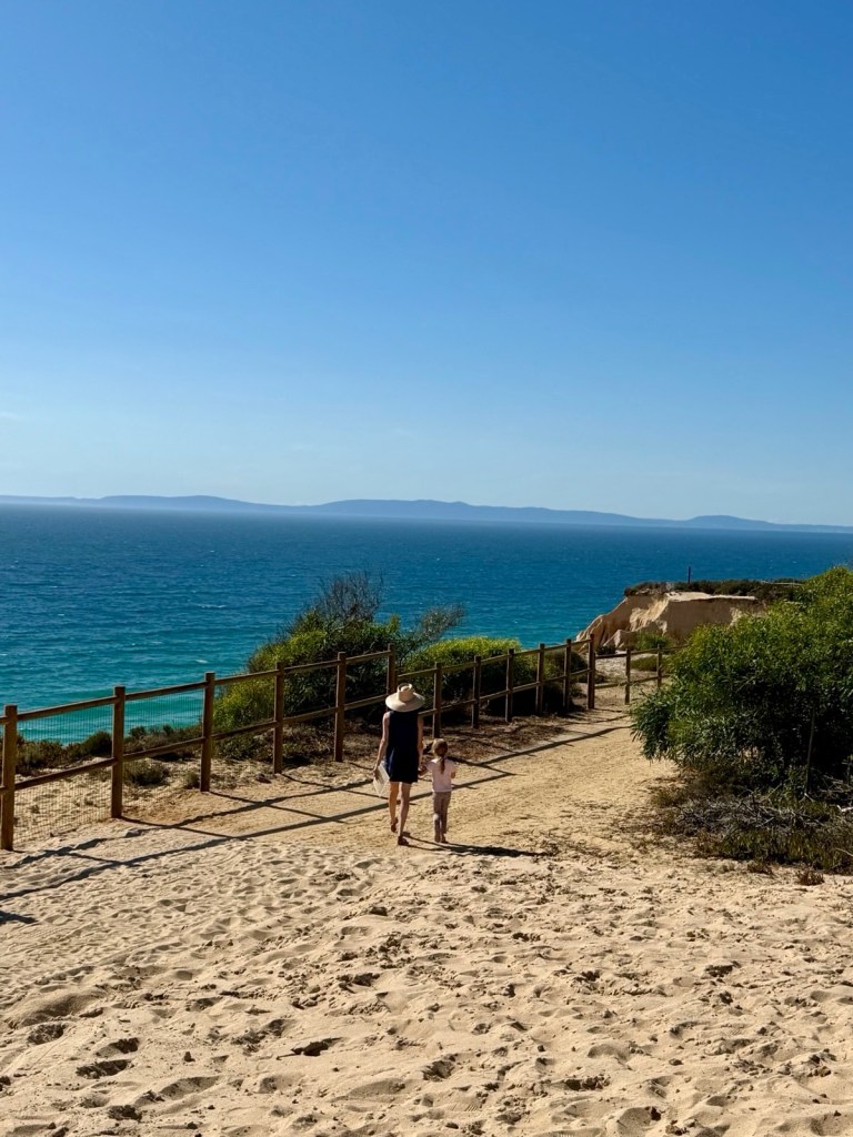 A woman in a sun hat and a young girl walk hand-in-hand down a sandy path overlooking the ocean.