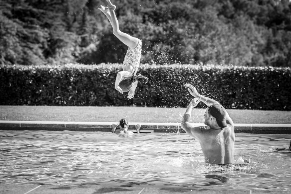 A child in a swimsuit doing a flip into a swimming pool with a person in a baseball cap reaching up towards them.