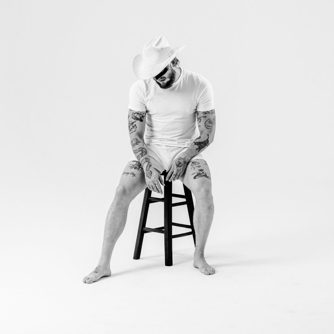 Orville Peck in a white t-shirt and briefs, wearing a white cowboy hat, sitting on a stool.