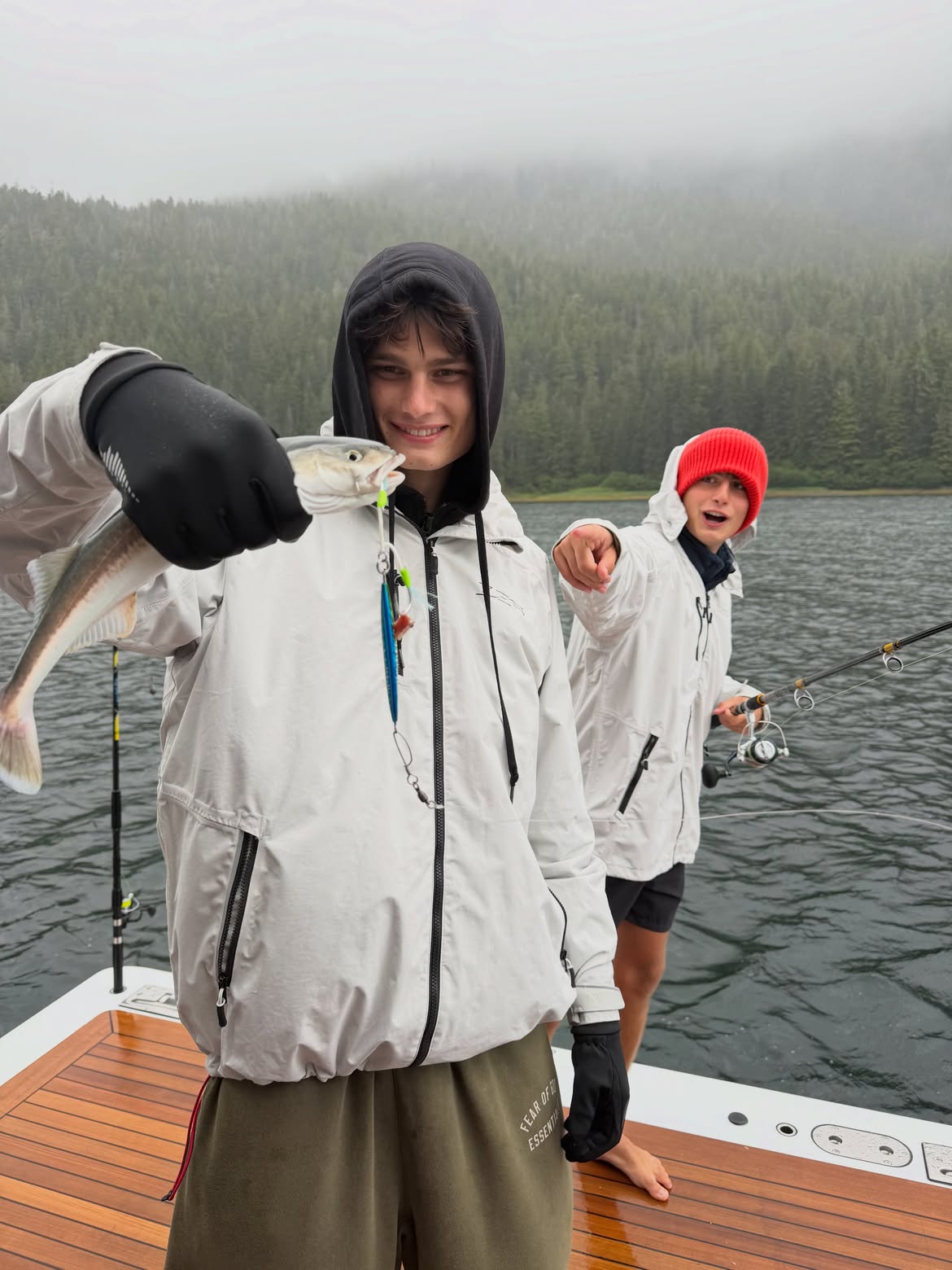 Two boys fishing on a boat, one holding up a fish and the other pointing.