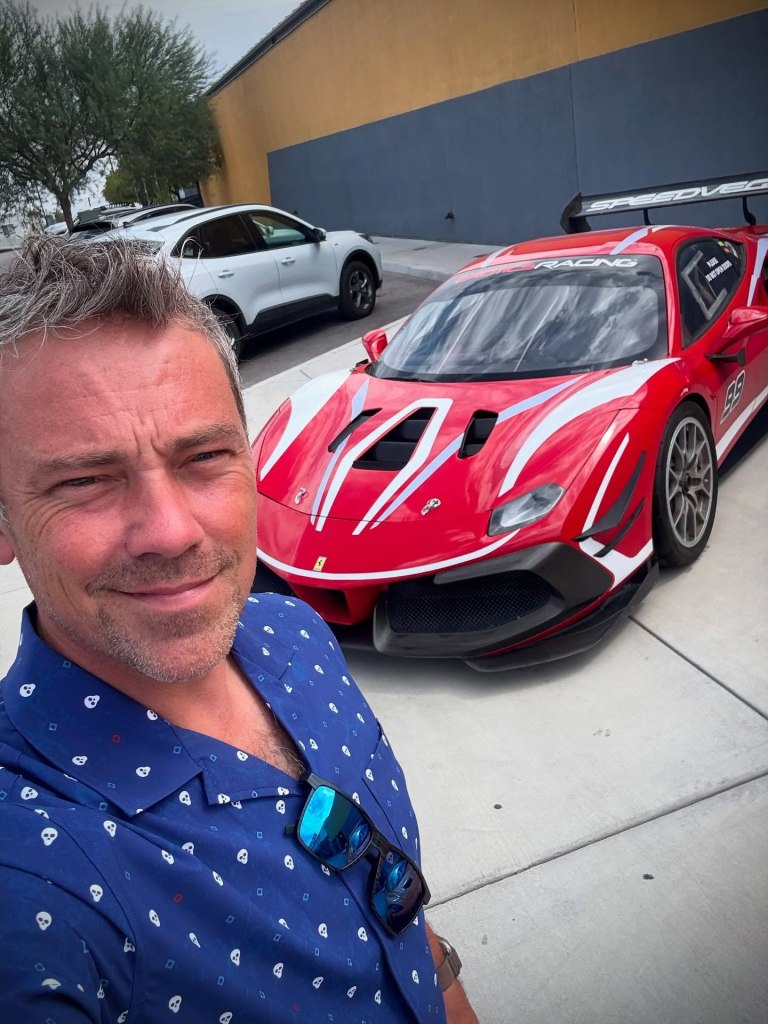 Kevin Baillie poses for a selfie with a red Ferrari race car.