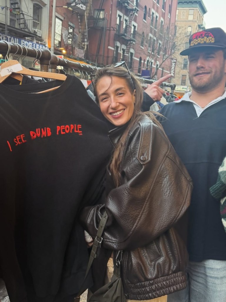 Amanda Batula holding up a black sweatshirt that says "I SEE DUMB PEOPLE" in red letters, with West Wilson pointing at her.