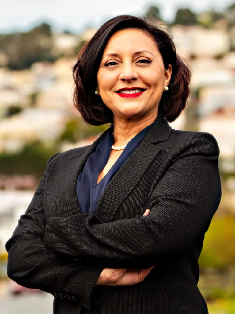 Myrna Melgar, a woman with dark hair and red lipstick, smiles while wearing a black blazer and pearl necklace.