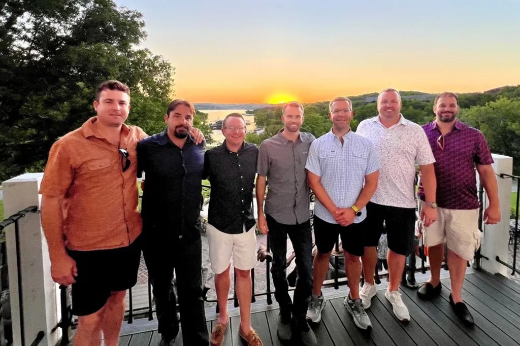 A group of six men standing together on a balcony with a sunset over a body of water in the background.