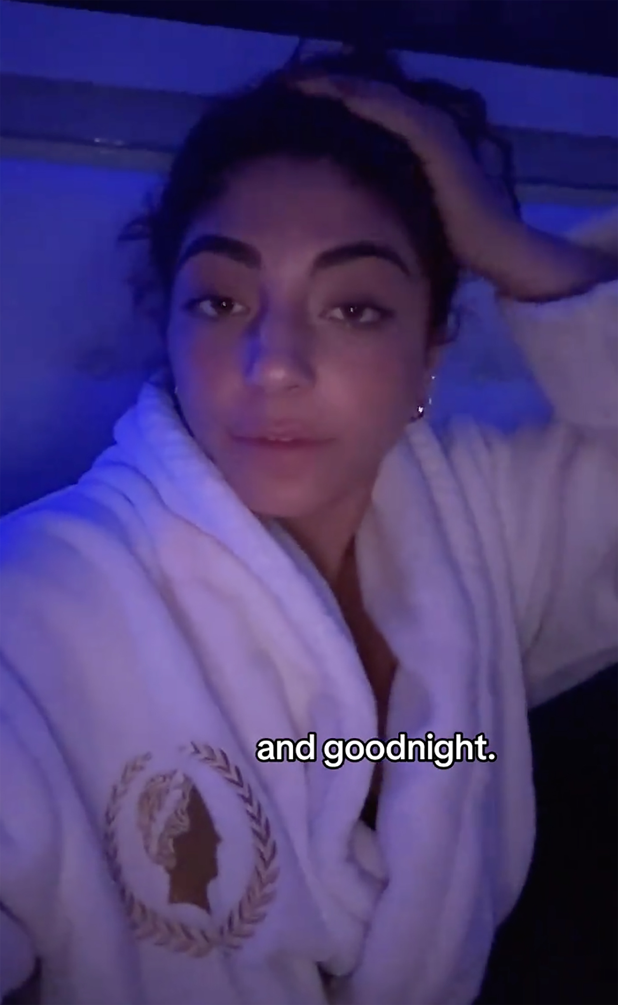 A woman wearing a white robe with an emblem of a woman's silhouette in a laurel wreath, with blue light shining on her face, and the words 