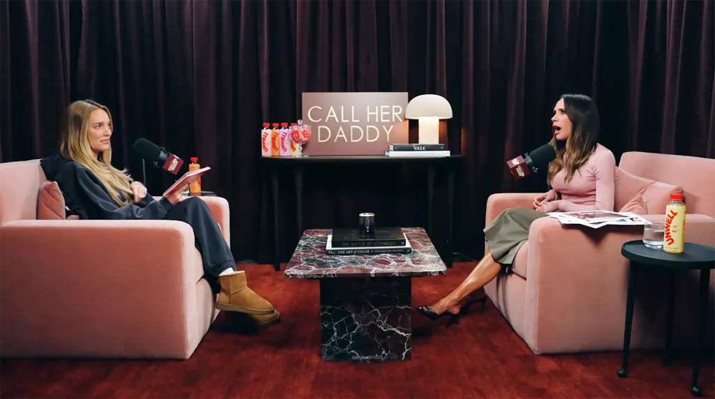 Victoria Beckham, in a pink top and grey skirt, being interviewed by Alexandra Cooper, in a black hooded top, black pants and brown boots, on the podcast "Call Her Daddy."