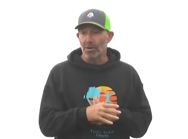 Man in a black hoodie and grey/lime green cap, speaking to the camera.