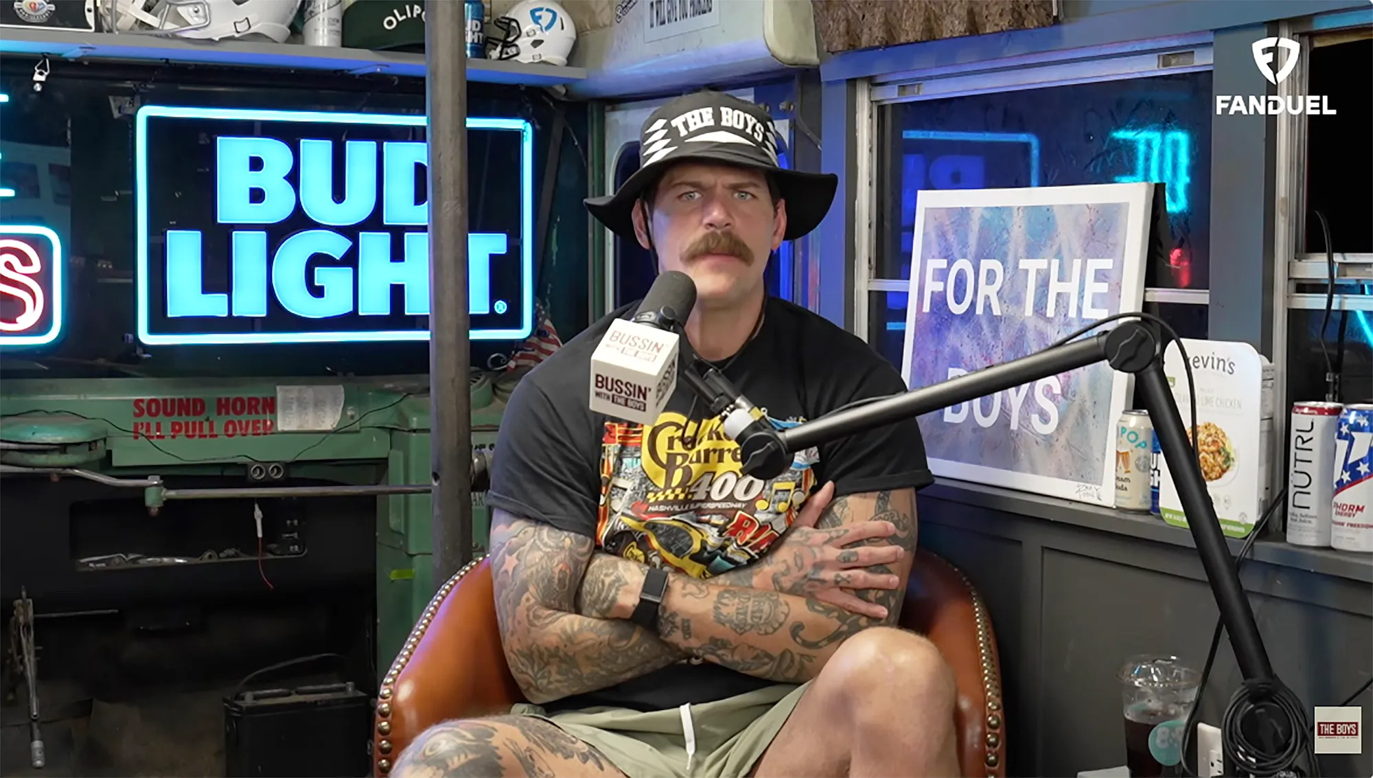 Taylor Lewan sitting in a chair with his arms crossed in front of a neon Bud Light sign.