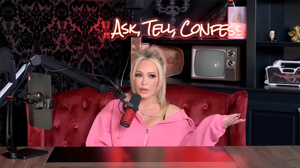 Bunnie XO sitting in a red armchair, speaking into a microphone with a neon "Ask, Tell, Confess" sign behind her.