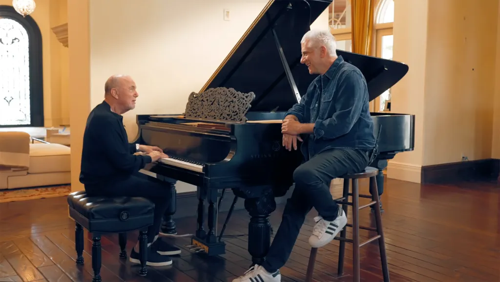 Billy Joel playing a grand piano while Rick Beato sits next to him.