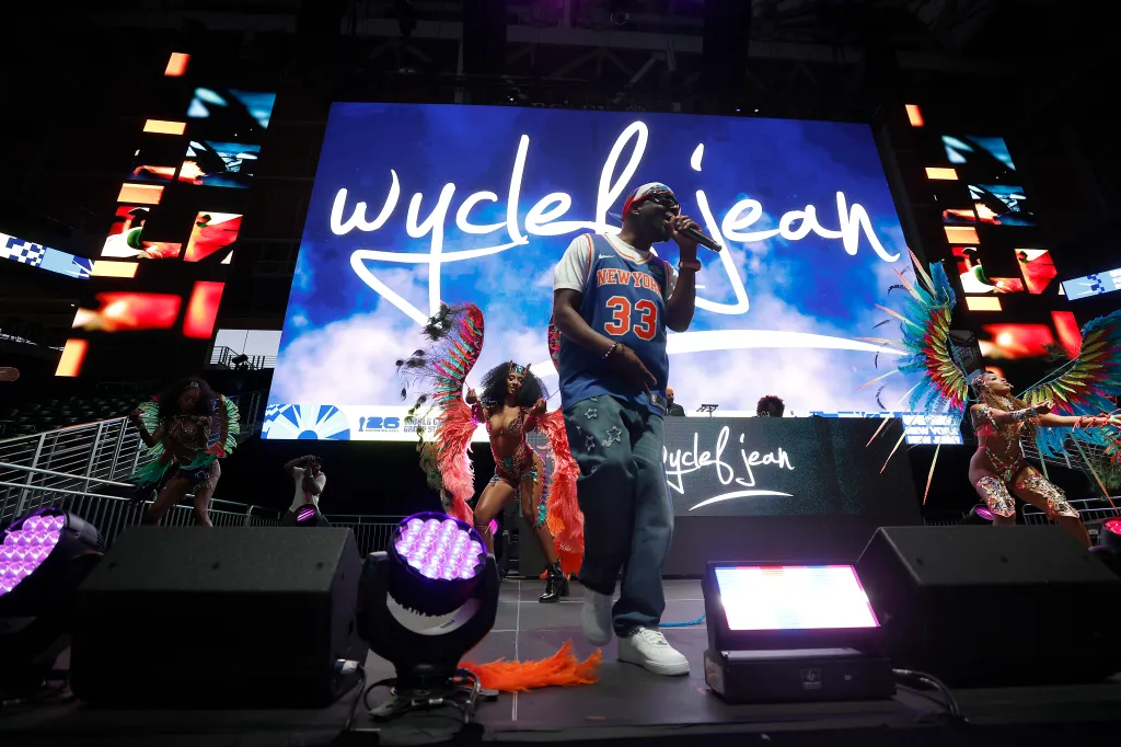 Wyclef Jean performs on stage in a "New York 33" jersey and jeans, with dancers in feathered costumes and a large screen displaying his name in the background.