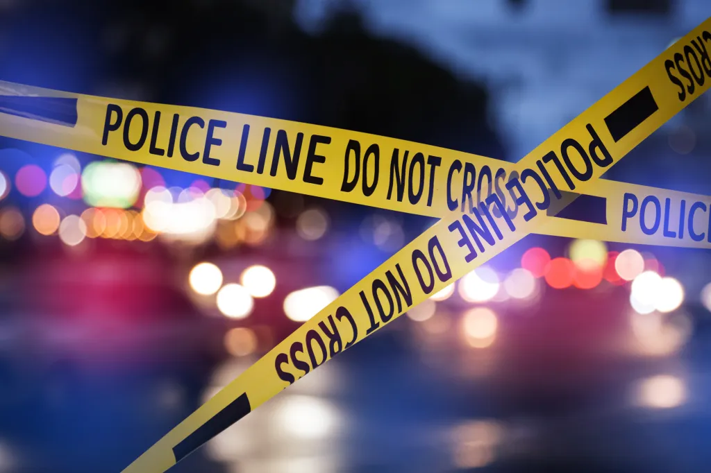 Yellow police tape reads "POLICE LINE DO NOT CROSS" with blurred red and blue lights in the background.