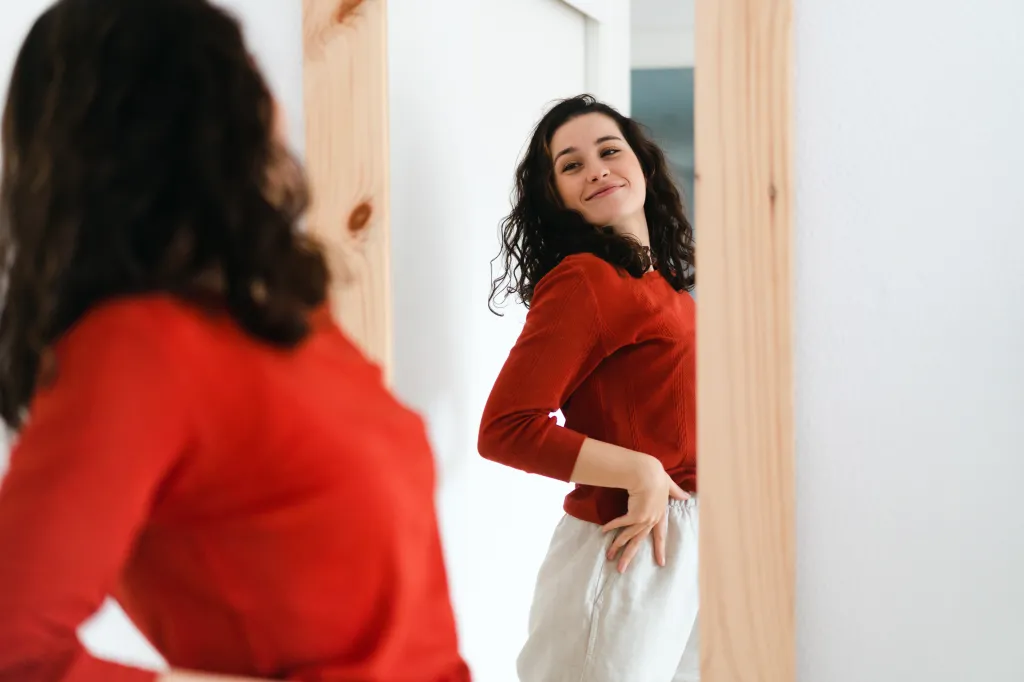 A young woman in a red sweater smiles confidently while looking at her reflection in a mirror.