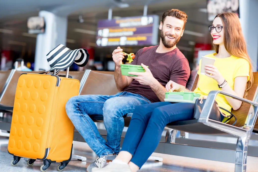 A happy couple eating snacks from lunchboxes in an airport waiting area with a yellow suitcase and striped hat beside them.
