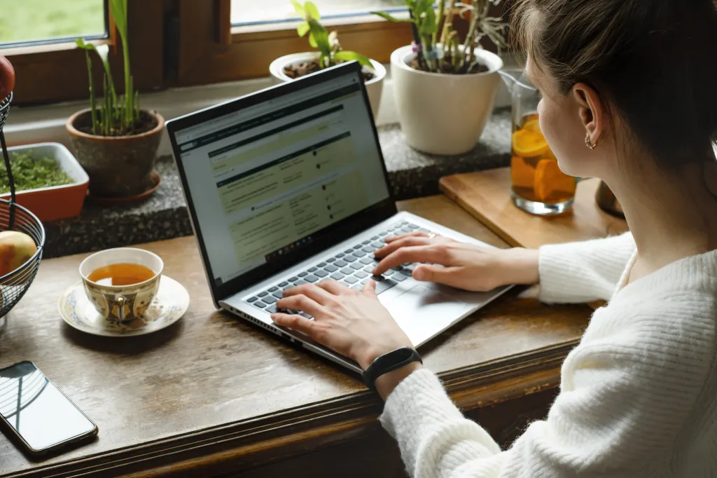 Woman working on a laptop at a table by a window at home.