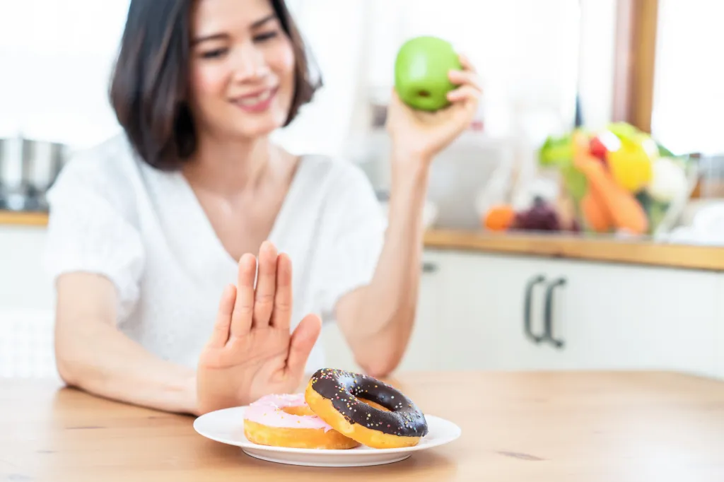 A woman pushes away donuts and chooses a green apple, making a healthy food choice.