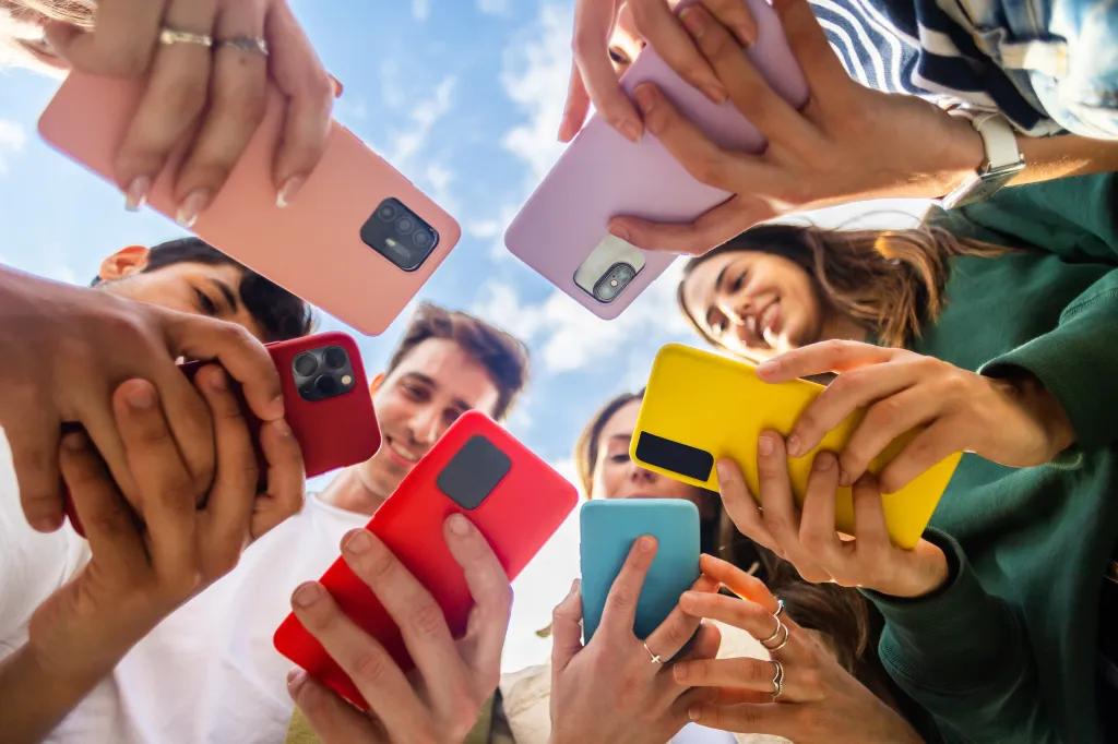 Low-angle view of a group of young people holding up their colorful smartphones, looking down at them.