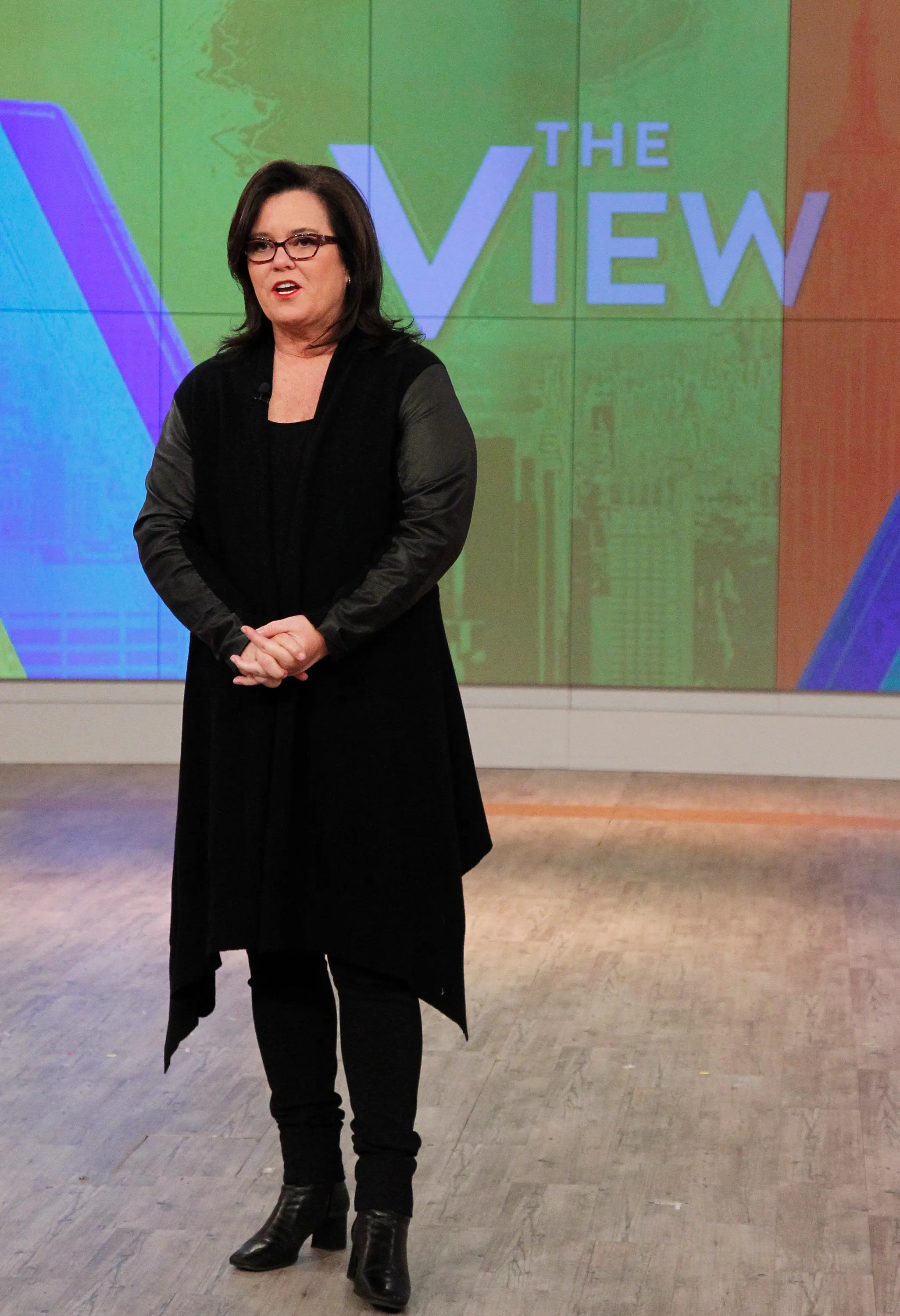 Rosie O'Donnell wearing a black long tunic with black boots and glasses, standing on the set of