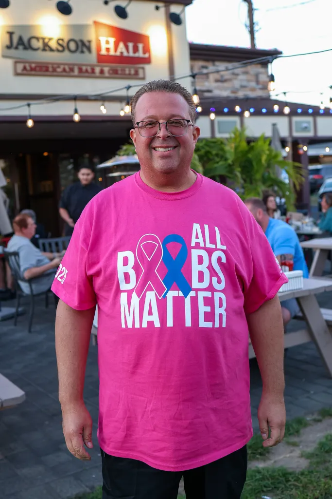 Michael Landesberg, a breast cancer survivor, smiles in front of his restaurant while wearing a pink "All Boobs Matter" shirt to promote a breast cancer fundraiser.