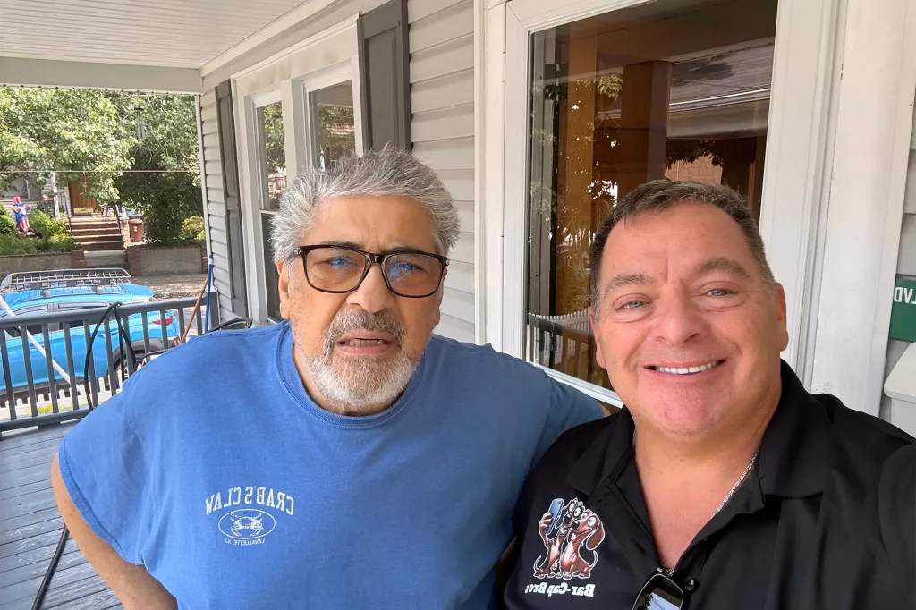 Two men posing on a porch, one in a blue t-shirt and the other in a black polo shirt.