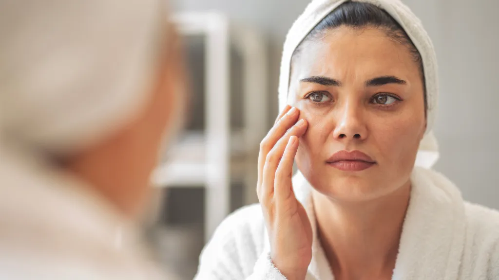 A woman with a towel wrapped around her head looking into a mirror and noticing wrinkles around her eyes.