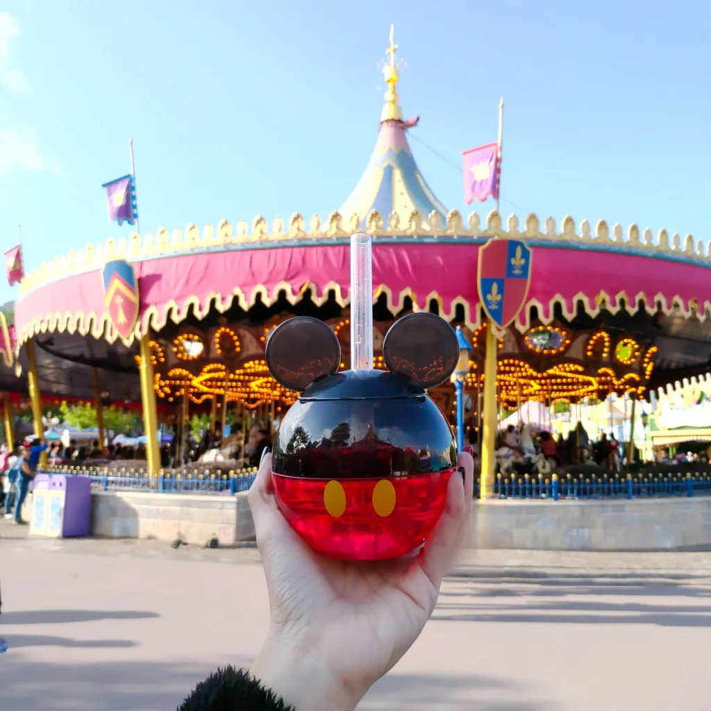 Hand holding a Mickey Mouse shaped souvenir drink bottle at a Disney theme park carousel attraction.
