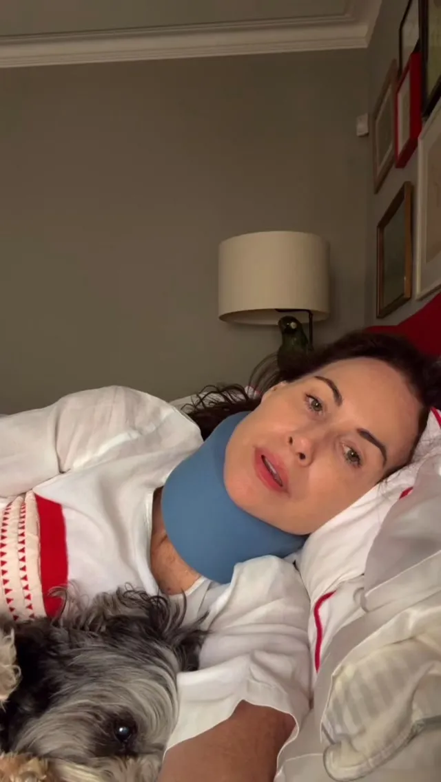 A woman in a blue neck brace lies in bed with her dog, while a green bird perches behind her.
