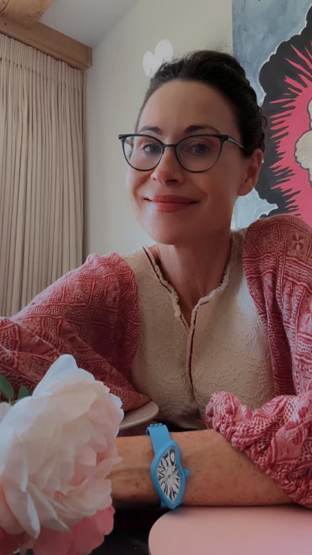 A woman wearing glasses and a patterned blouse smiles at the camera, with a blue melting clock-style watch on her wrist and a large pink flower in the foreground.