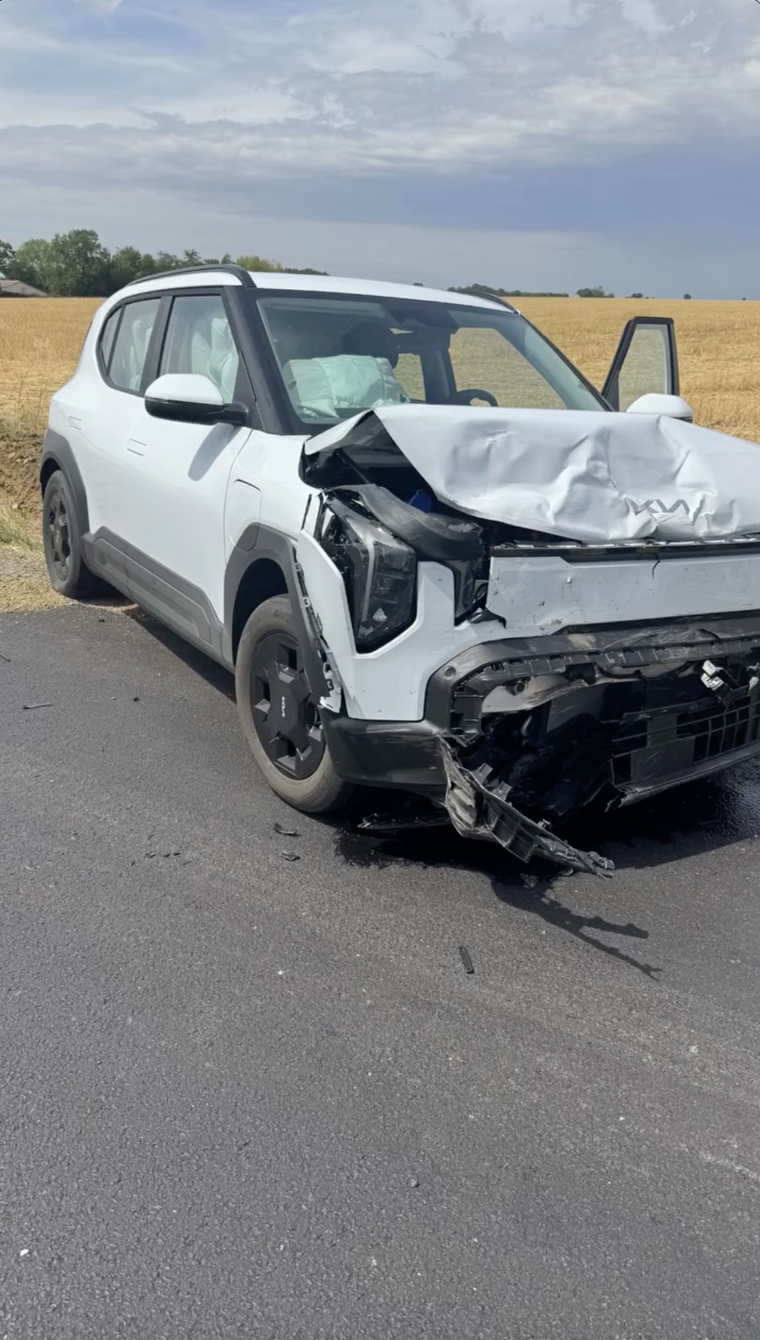 A white car with significant front-end damage and deployed airbags, parked on the side of a road next to a field.
