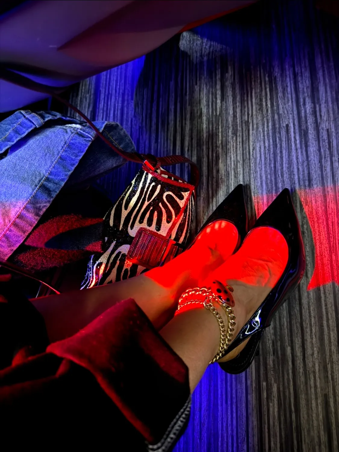 Person's feet wearing black high heels and a gold chain ankle bracelet, with a zebra print handbag and denim jacket, under red and blue lighting.