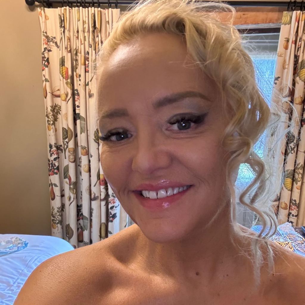 A woman with blonde hair, dark eyeliner, and shiny lips smiles brightly.
