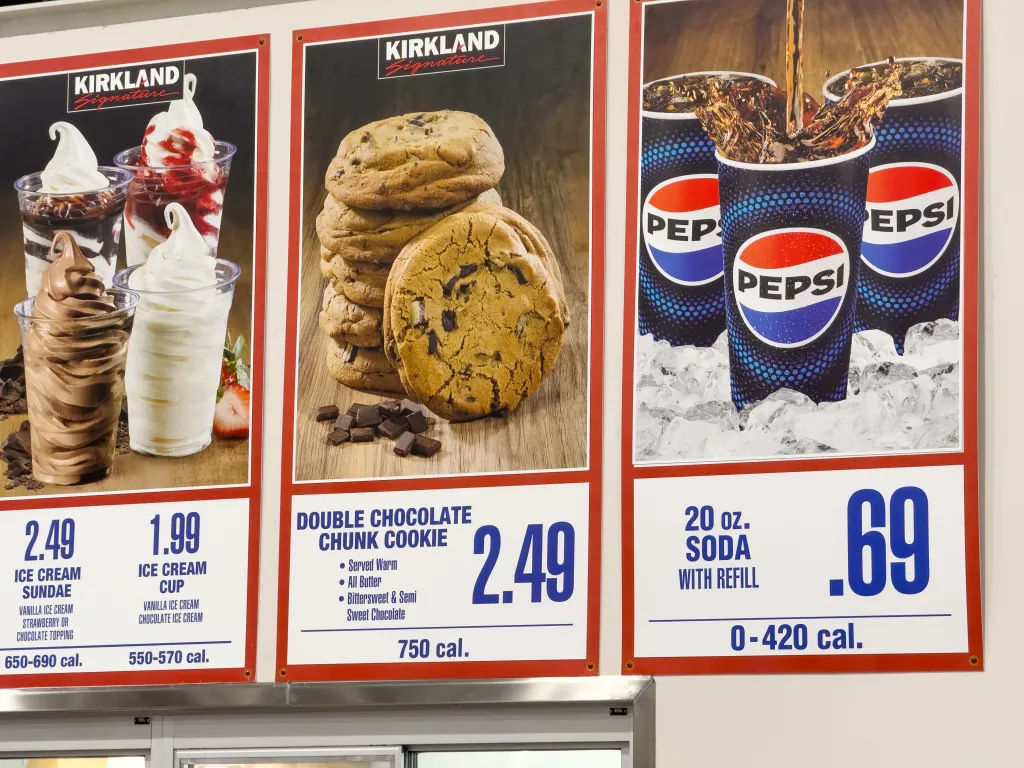 Costco food court menu signs showing Kirkland Signature ice cream sundaes and cups, a double chocolate chunk cookie, and 20 oz. soda with refill.