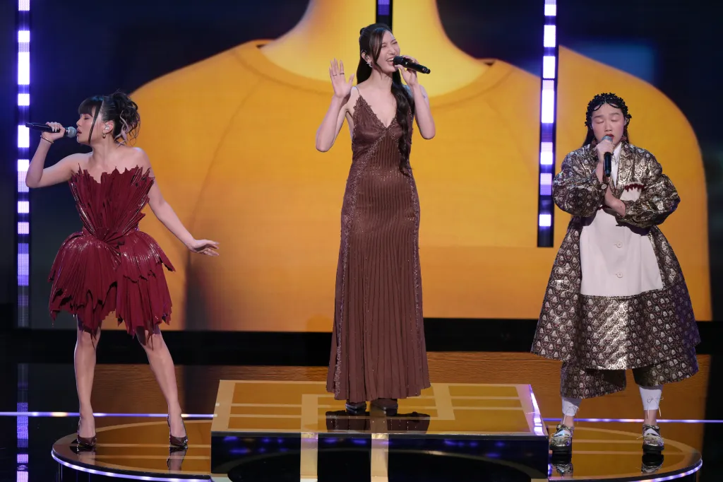 Three women singing on stage, one in a red dress, one in a brown dress, and one in a gold and white coat.
