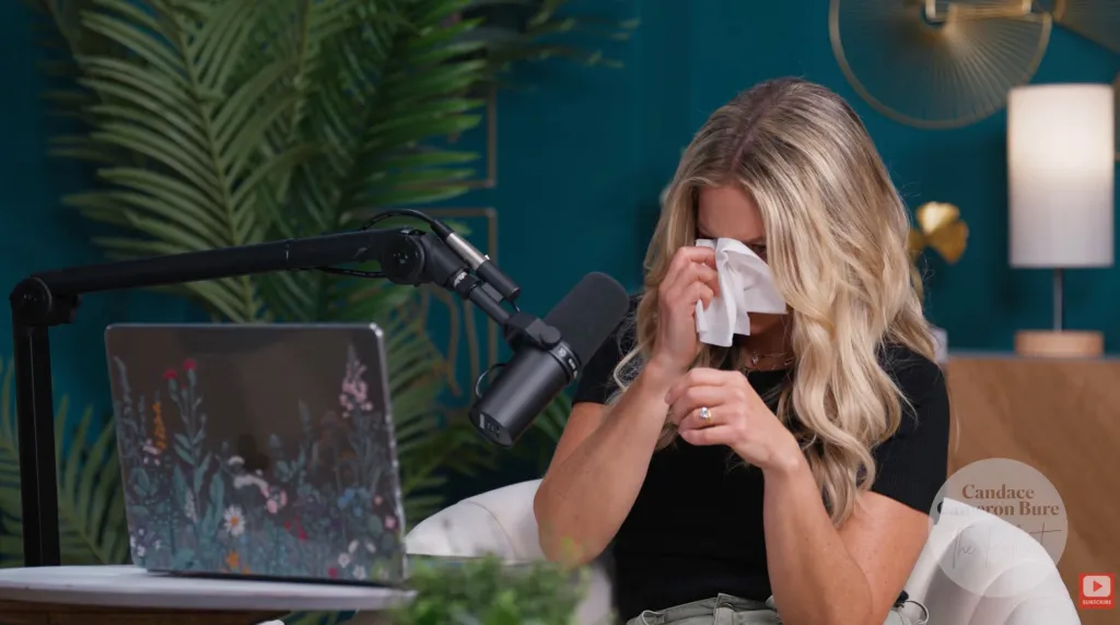 A blonde woman sitting at a desk with a microphone and laptop, dabbing her eyes with a tissue.