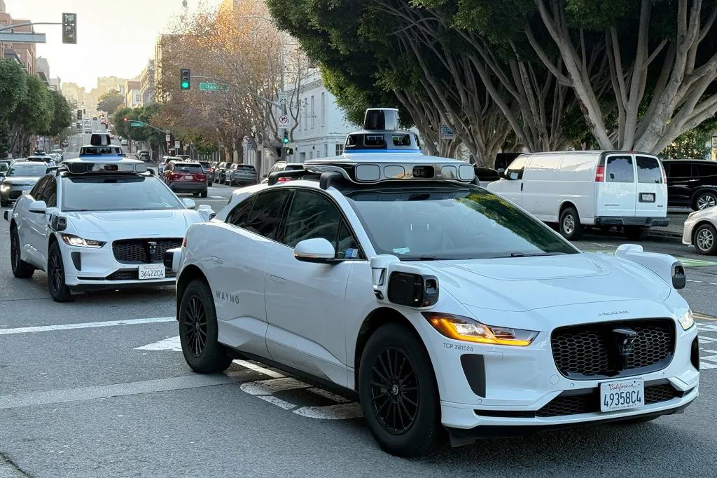 A line of white Waymo self-driving Jaguar I-Pace electric cars with sensors on their roofs and fenders driving on a city street.