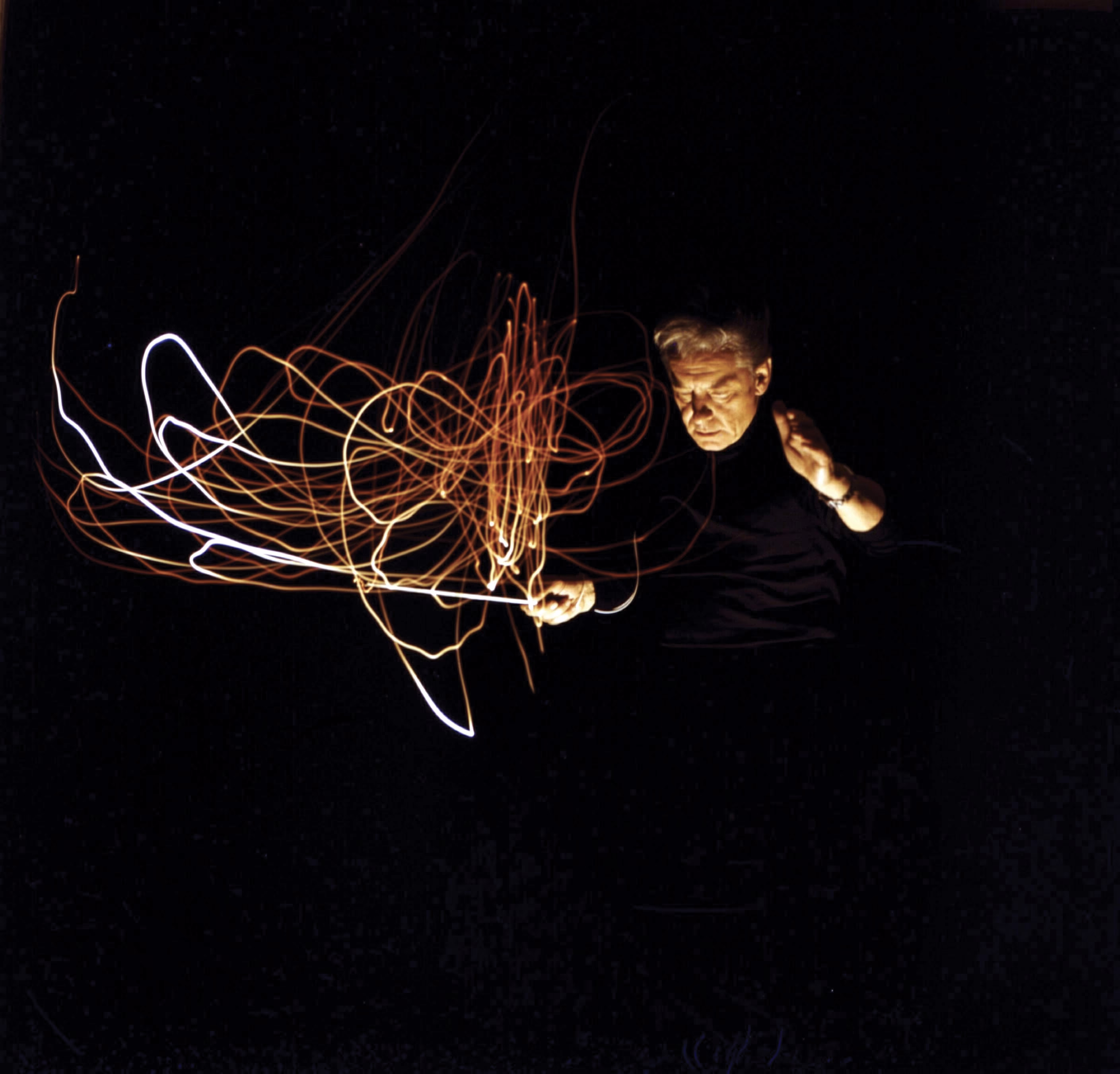 color time-lapse photo of man strongly lit from below, where the baton's movements appear as multiple tangled trails of reflected light