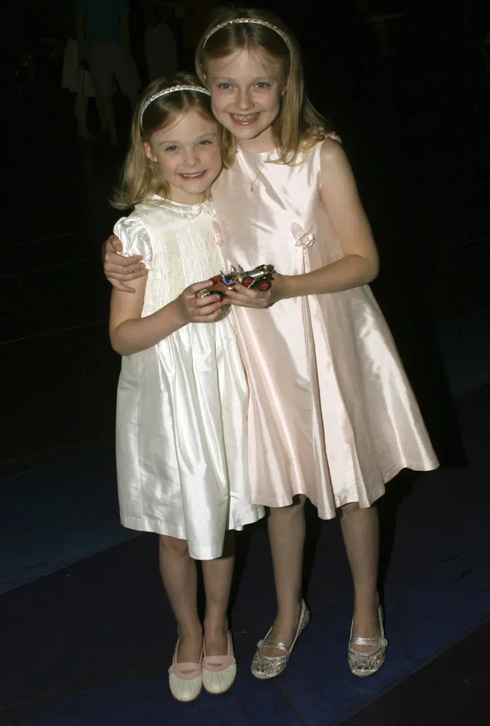 Sisters Elle and Dakota Fanning, both wearing shiny dresses and headbands, pose with their arms around each other.