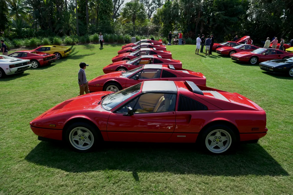 A young boy walks past a display of red 1980s Ferrari 328 GTS sports cars parked on green grass.