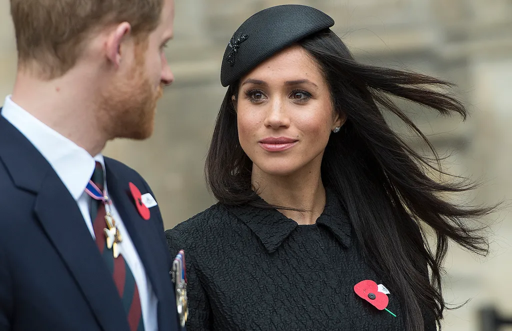 Meghan Markle wearing a black fascinator and textured black coat, with a red poppy pinned to her lapel, looks toward Prince Harry.
