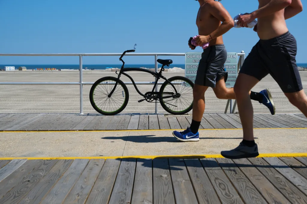 Two shirtless men run along the boardwalk overlooking the beach, in Wildwood, New Jersey. A bicycle is parked next to them.